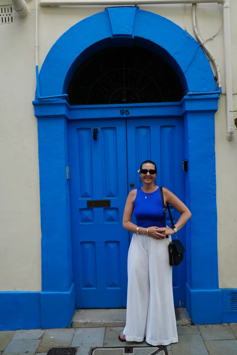 Heidi in a blue top and white wide-leg trousers in front of a blue Georgian doorway in Gibraltar