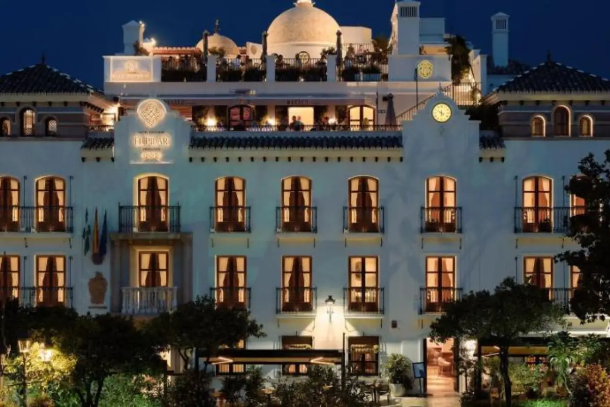 The edifice of Hotel Silken el Pilar in Estepona at night with people on the balcony and the lights on in the windows.