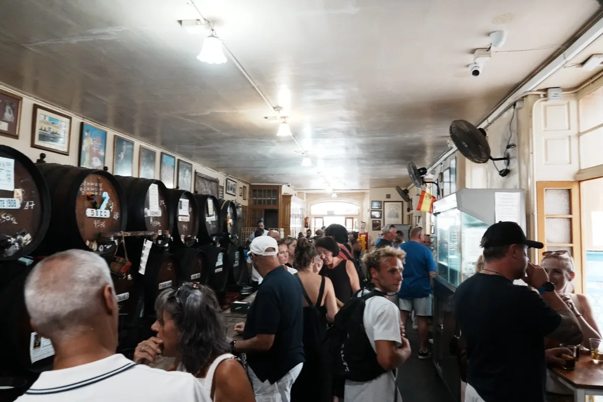 A crowd standing shoulder to shoulder along the long bar at Antigua Casa de Guardia, wine barrels and framed photographs down the wall behind them