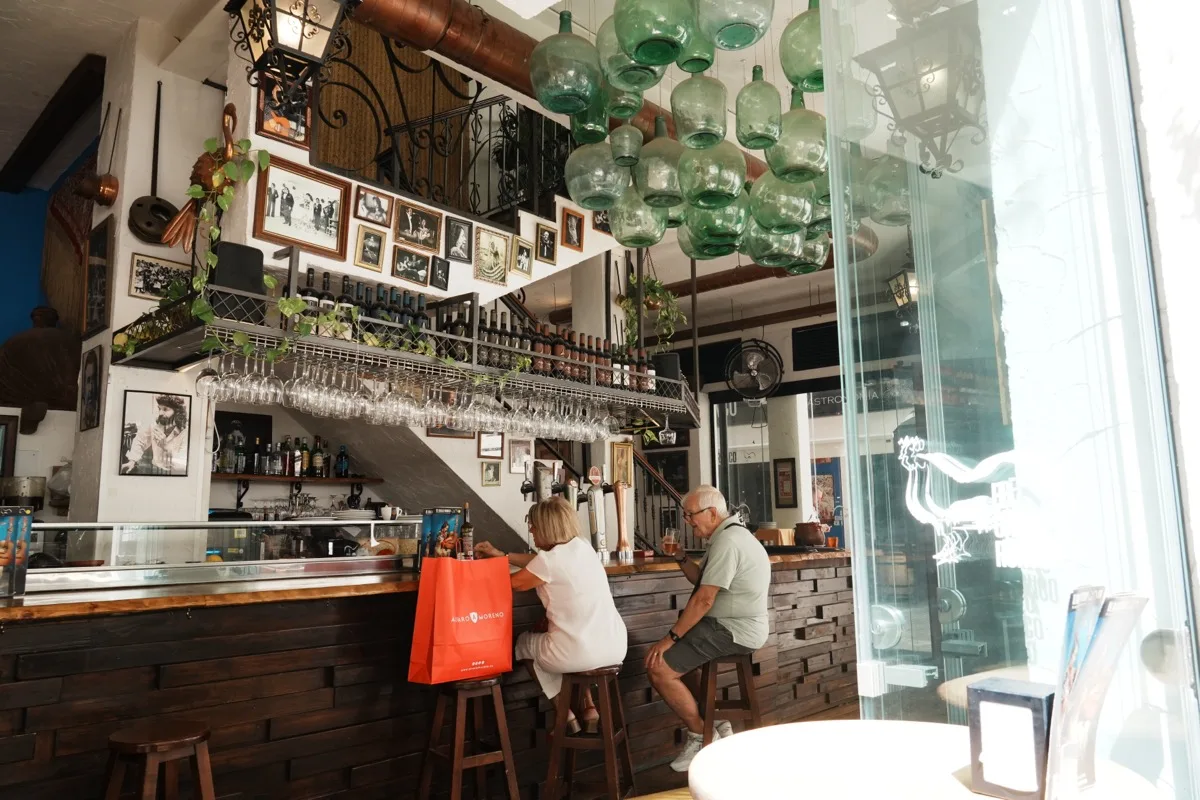 The bar inside El Gallo Ronco, green glass demijohns hanging from the ceiling, bottles and glasses on the shelf above the counter and two customers on stools by the window