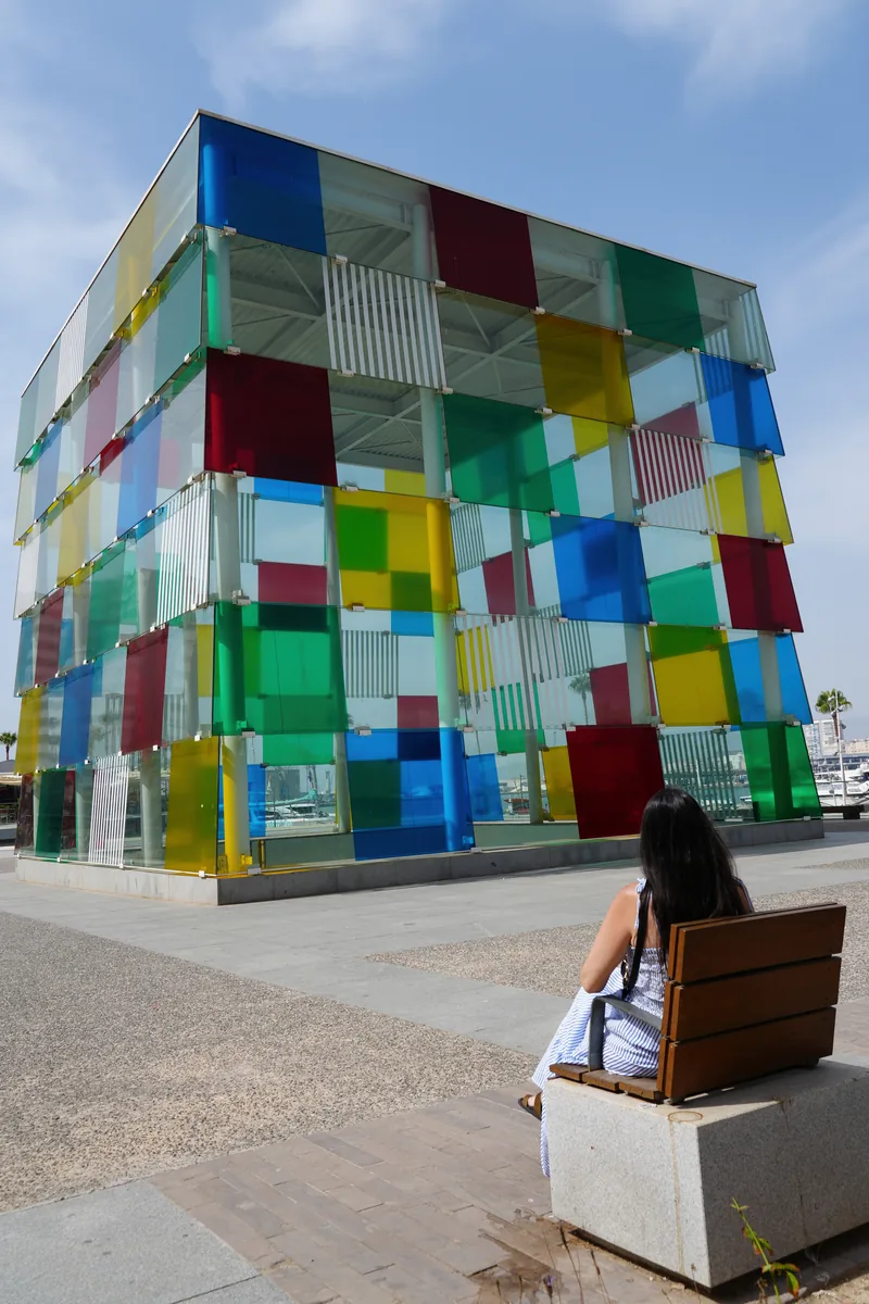 Heidi sitting on a bench in front of the colourful glass Cube of Centre Pompidou Malaga