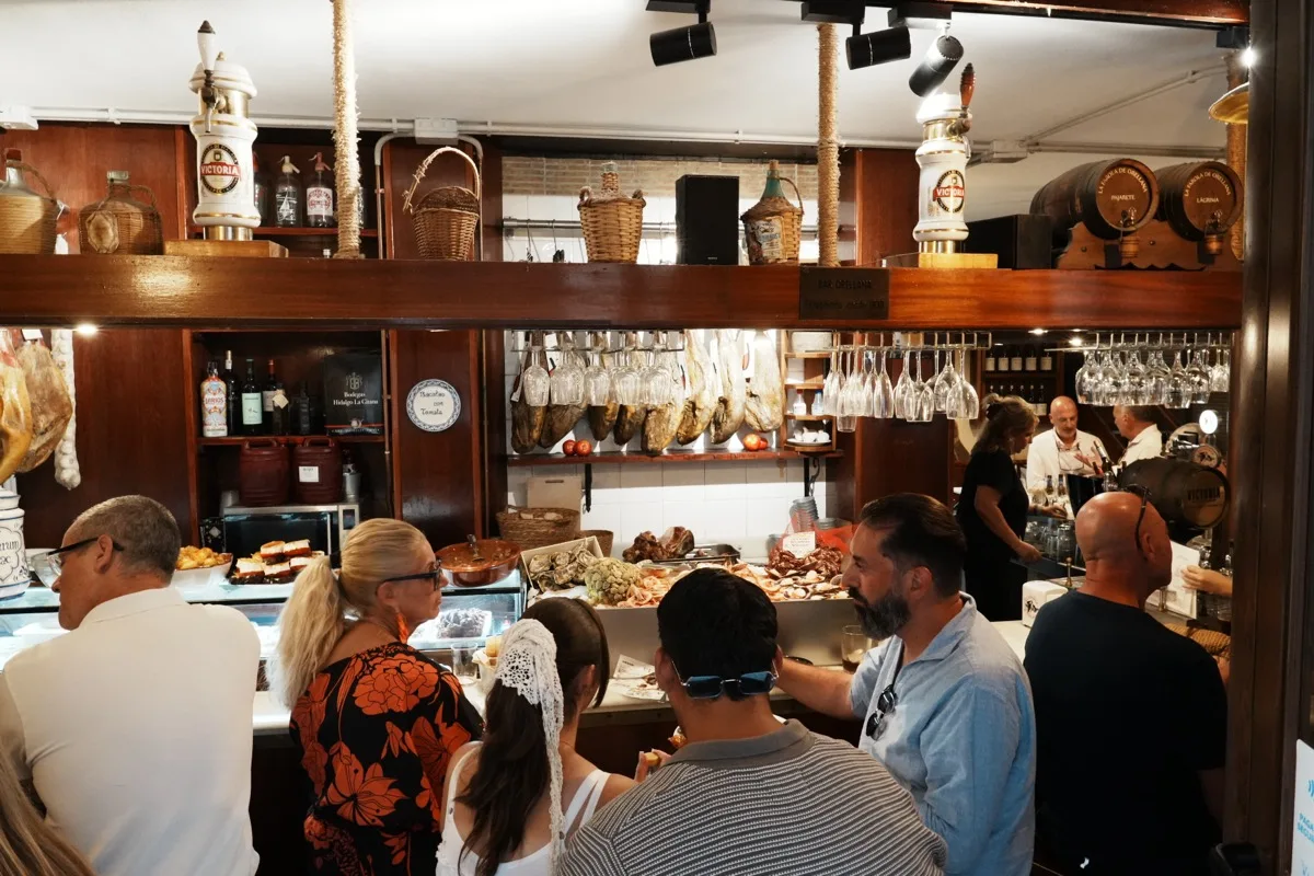 Customers crowded at the counter of La Farola de Orellana, platters of food laid out under hanging hams and racks of glasses