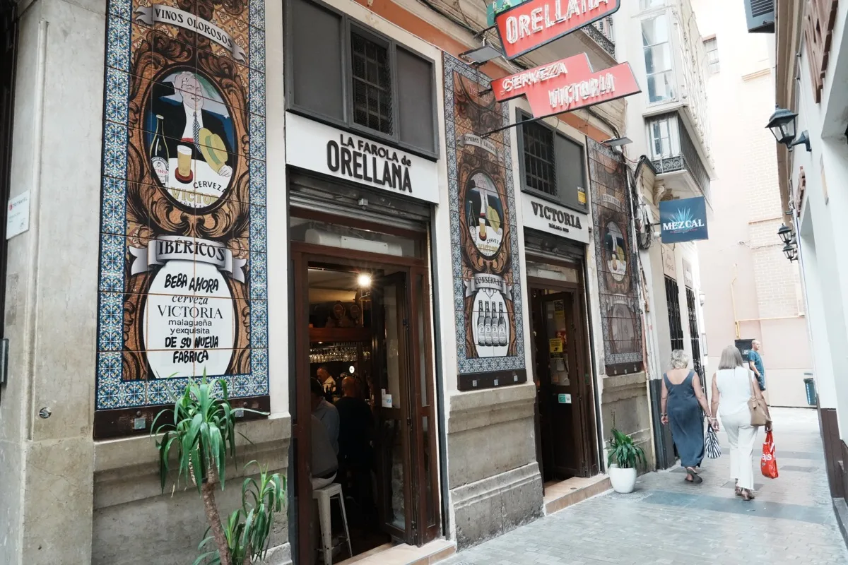 The tiled frontage of La Farola de Orellana on narrow Calle Moreno Monroy, with its painted murals, a Victoria beer sign and people walking past the door