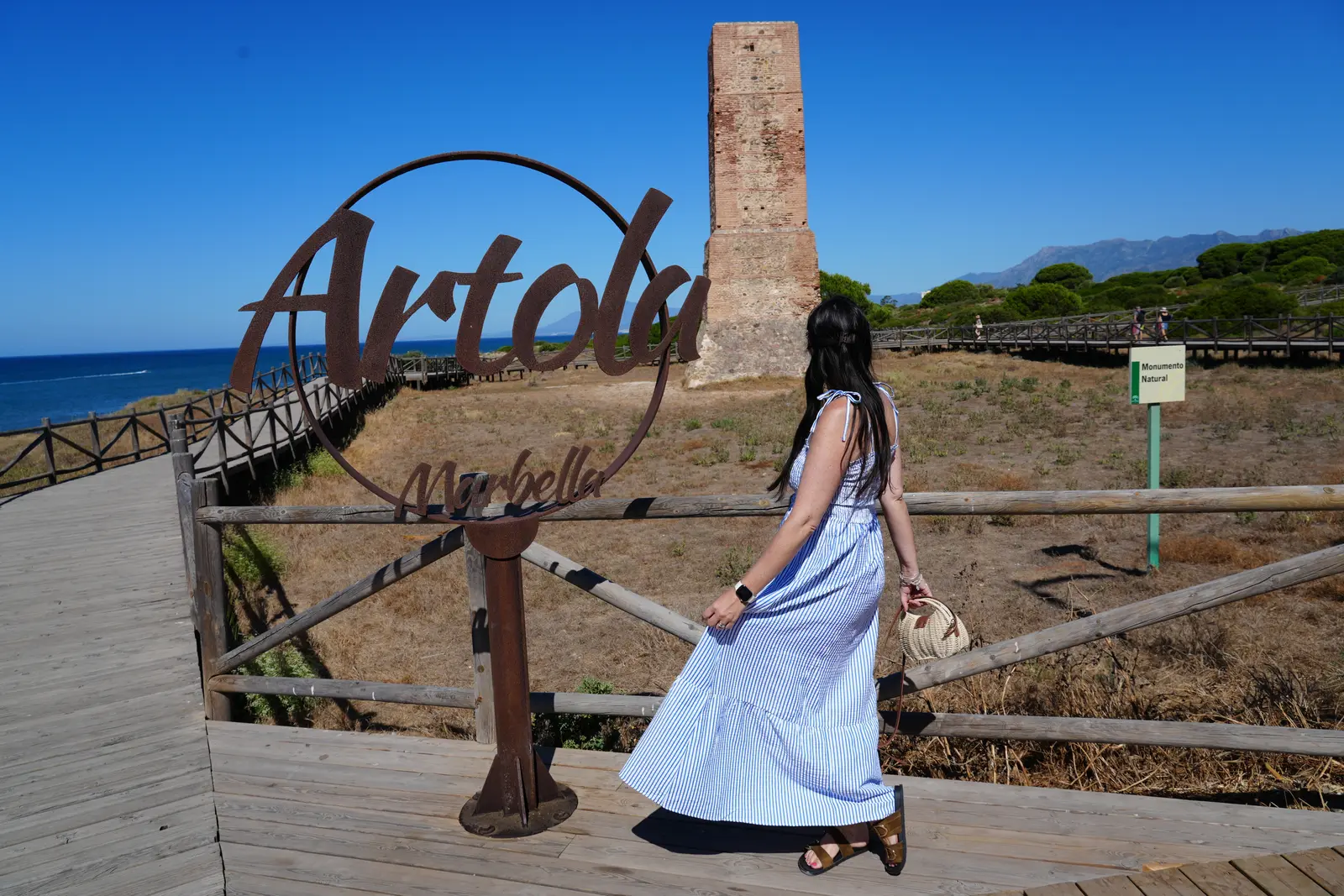 A woman standing in front of the Artola sign at Artola Marbella Beach with the large brick tower behind her.